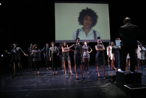 A group of people are on a stage in front of microphones. There is a projection behind them of a woman. A man is on a pedestal facing them with a laptop in front of him.