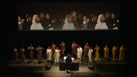 A group of people are on a stage in front of microphones. There is a projection behind them of three screens of people. A man is standing behind a table, facing them with laptops in front of him.