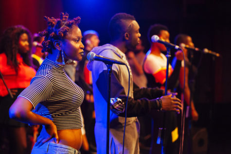 Men and women are standing on a stage, facing forward, in front of microphones.
