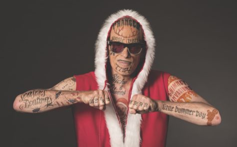 A man is pictured from the waist up looking at the camera. He is wearing a red shirt with a hood that is lined with white fur. He is covered in various tattoos and is wearing sunglasses.