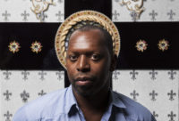 A man standing in front of a wall with white wallpaper with silver crosses on it. Behind him is a large, black gross with small gold designs and a round gold design is immediately behind his head. He is looking at the camera and is wearing a blue, button-down shirt.