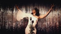 A woman in a white gown stands at a microphone on a brightly lit stage in front of a shimmering silver backdrop, with her arms outstretched, chin raised, and eyes closed.