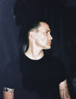 A man with short, dark hair and wearing a black t-shirt, looks towards the right of the frame.
