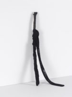 A piece of wood leaning on a white wall with a black nylon stocking attached to it.