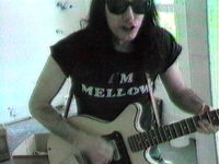 Man with long dark hair, sunglasses, and a t-shirt that says, “I’m mellow” plays a cream-colored electric guitar.