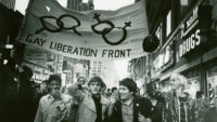 A black and white film still of people marching down a city street below a banner that says, "Gay Liberation Front".