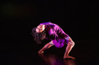 Dancer in purple flowing shirt and knee pads bends far backward with her head toward the audience.