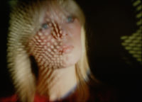 A film still of a person who is slightly blurred. There is a reflection on their face of many dots.