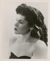 A black and white publicity photo of a person in a dark dress with long, dark hair, wearing makeup and looking off to the left of the frame.