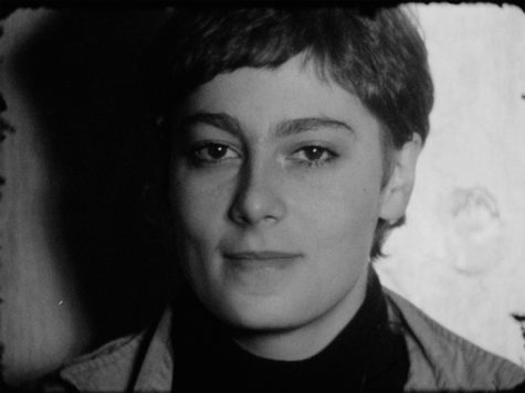 Black and white film still of a person's face. They are looking at the camera and smiling.
