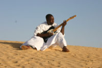 A person dressed in white, sits outside on sand in the daylight while playing guitar.