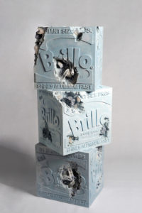 A sculpture depicting three stacked gray Brillo boxes with various large holes in them. Some of the holes have clear crystals in them.