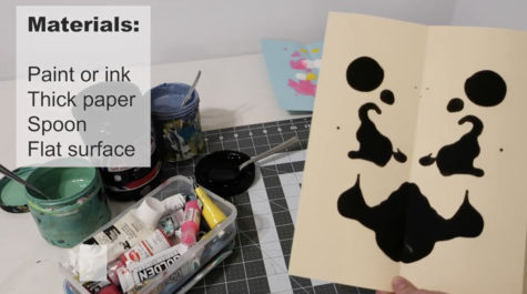 A still from a video of a table with art supplies with someone holding a Rorschach painting in black paint on creme paper in the foreground. A materials list is on the left side.