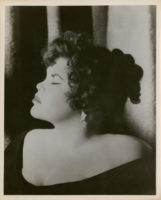 Black and white photograph of a person with dark hair and wearing a dark dress and earrings. Their eyes are closed and they are looking off to the left of the frame.