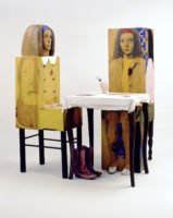 3D sculpture of two people wearing yellow sitting down on chairs at a table.