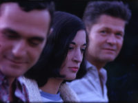 Color film still of three people sitting side-by-side. The person in the back is looking at the camera. The other two are looking in other directions. The first person is smiling while looking down.