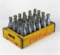 A yellow wood crate with "Drink Coca-Cola in bottles" and "Have a Coke" in red on each side. The crate has 23 glass Coke bottles in it that are spray painted silver.