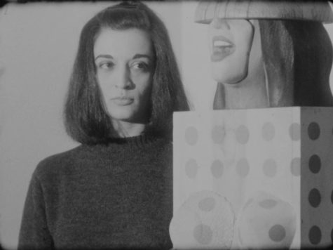 Black and white film still of Marisol Escobar looking at one of her sculptures that she is standing next to.