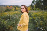 A person with red hair wearing a yellow jacket stands in a field of green grass. They are looking towards the camera.