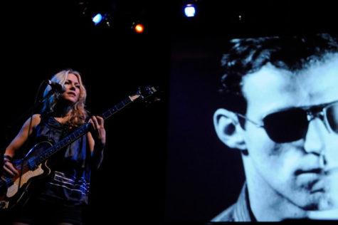 A person plays a bass behind a microphone on a stage as a black and white film is playing behind them.