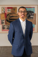A smiling person in a blue dress coat and white button up shirt poses in a gallery in front of an artwork by Jean-Michel Basquat and Andy Warhol. The person is wearing glasses and has a goatee.