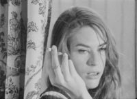 A black and white film still of a person holding a cigarette in their hand which is next to the right side of their face. They are looking off to the right side of the frame.