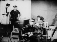 Black and white film still of a band playing to the right of the frame while a policeman stands in front of a white backdrop on the left side of the frane.