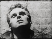 Black and white film still of a person from the neck up standing in front of a stone wall with their head tilted back and their eyes closed.