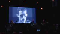 Video still of a band playing on a stage as a film by Andy Warhol plays in the background.