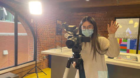 A person behind a camera is wearing a mask while looking and waving towards the person taking the photo.