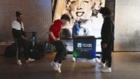 Two people stand while one dances in front of a DJ stand that says "Tech 25" in the entrance space of The Andy Warhol Museum.
