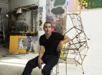 A person is sitting in their artists studio on one of their sculptures made up of any metal chairs.