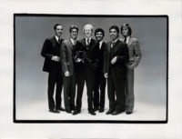 Black and white photograph of Fred Hughes, Vincent Fremont, Andy Warhol, Ronnie Cutrone, Bob Colacello, Sean Byrnes, and dog Archie held by Warhol. The men are all wearing suits and ties and standing in front of a gray background. They are all smiling and facing forward.