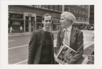 Black and white photograph of John Waters and Andy Warhol standing on a sidewalk on Madison Avenue in New York. Warhol is looking towards Waters and smiling while holding Interview magazines in his hand. There is a large car behind them and stores across the street in the background.