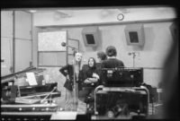 Black and white photograph of three of the members of the Velvet Underground with Nico in a recording studio singing in front of a microphone.