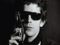 Black and white film still of Lou Reed closeup wearing dark sunglasses and holding a glass bottle of Coke next to the right side of his face.