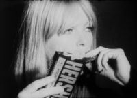 Black and white film still of Nico looking towards her left holding a Hershey bar with her right hand in front of the right side of her face. She is eating a piece of chocolate.