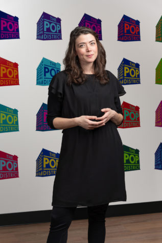 Ani Martinez, who is wearing a black dress, stands in front of a wall covered with The Pop District logos in various colors.