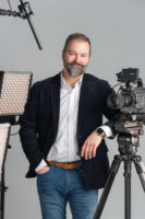 Christian Lockerman, who is wearing jeans, a white button up shirt, and black suit coat, is standing in front of a gray background while leaning on a video camera on a tripod with his left elbow. To his right is a lighting set up.