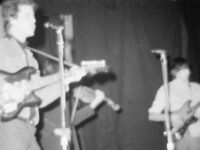 Blurry black and white film still of a band playing on a stage.