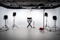 An empty studio with white walls, a directors chair, and various lighting equipment all around.