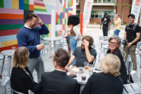 A group of people gathered outside on Silver Street, the alley behind The Andy Warhol Museum, that includes a mural painted by the artist, Typoe, made up of various colorful shapes on the wall of the museum in the background. Some are standing and some are sitting at a table.