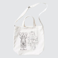 White tote bag featuring a drawing of a cartoon-like figure by the artist KAWS and a robot by Andy Warhol, both in a playful design. The bag has a white strap and a plain background.