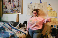 Izsys Archer is wearing a pink sweatshirt with graphic designs, jeans, and sunglasses, and is extending one arm towards a canvas on an easel standing in a studio surrounded by various artworks, including sketches and colorful paintings.