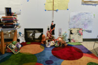 An artwork of an individual knelt on a colorful rug, surrounded by stacks of books, papers, and various artistic supplies. Some dolls and crafted figures are scattered around the individual. A world map and other artworks hang on the wall behind.