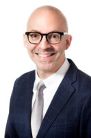 Portrait of Mario R. Rossero smiling, wearing glasses and a dark suit, set against a plain white background.