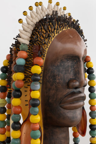 Close-up of a sculptural artwork depicting a face with ornate, colorful bead necklaces and a detailed headdress, incorporating elements such as small spheres and spikes.