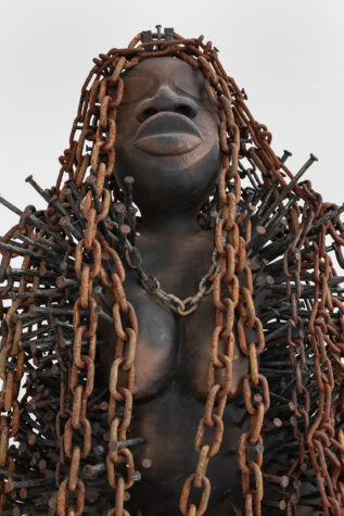 Sculpture of a figure with eyes closed, covered in cascading rusty chains, against a plain background.