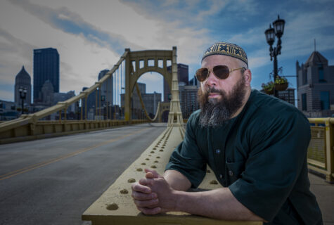 Sharif Bey, wearing sunglasses and a patterned cap, stands on the Andy Warhol Bridge overlooking the cityscape of Pittsburgh under a partly cloudy sky.