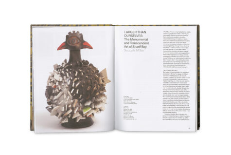 An open book displaying an article titled "Larger Than Ourselves: The Monumental and Transcendent Art of Sharif Bey" by Sequoia Miller. The left page shows a photograph of a sculptural artwork resembling a bird with a textured, spiky body. The right page contains text about the artwork.